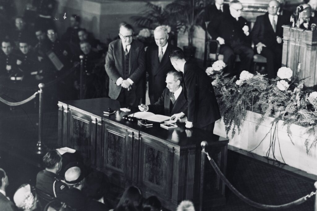 4 de abril de 1949, o secretário de estado dos EUA, Dean Acheson, assina o tratado da OTAN. A esquerda, na foto, o vice-presidente Alben W. Barkley conversa com o presidente Harry Truman. NATO Photo