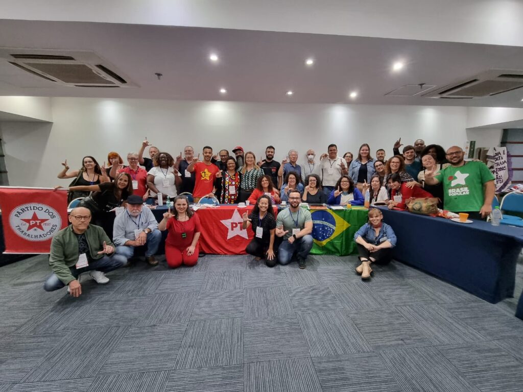 Uma grande reunião de militantes e apoiadores do Partido dos Trabalhadores (PT) e dos movimentos populares. O grupo, composto por homens e mulheres de diferentes idades e origens, posa sorrindo para a foto em uma sala ampla. Algumas pessoas usam camisetas vermelhas com símbolos do PT, enquanto outras vestem roupas casuais. À frente da mesa, bandeiras do Brasil e do PT estão estendidas, reforçando o engajamento político e a luta coletiva. Muitos participantes fazem o gesto de "L" com as mãos, simbolizando o apoio ao ex-presidente Lula e às causas progressistas.