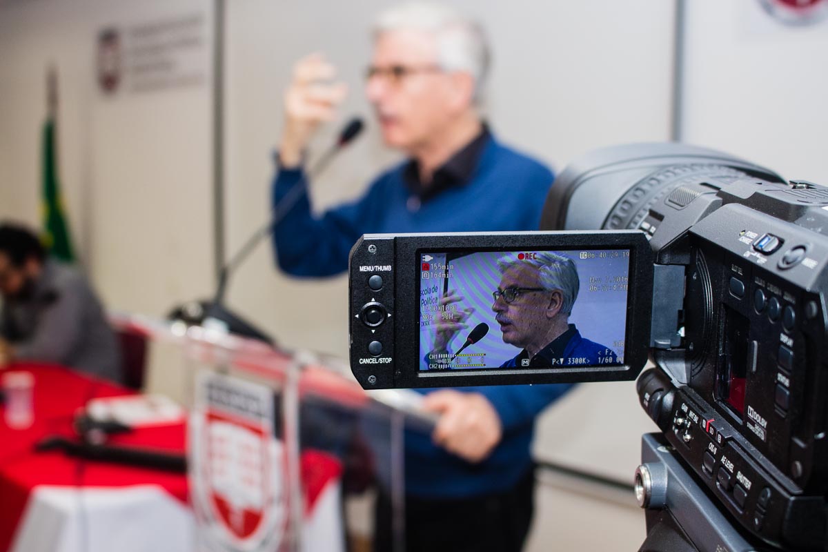 Homem palestrante gravado em evento, tela de câmera em primeiro plano.