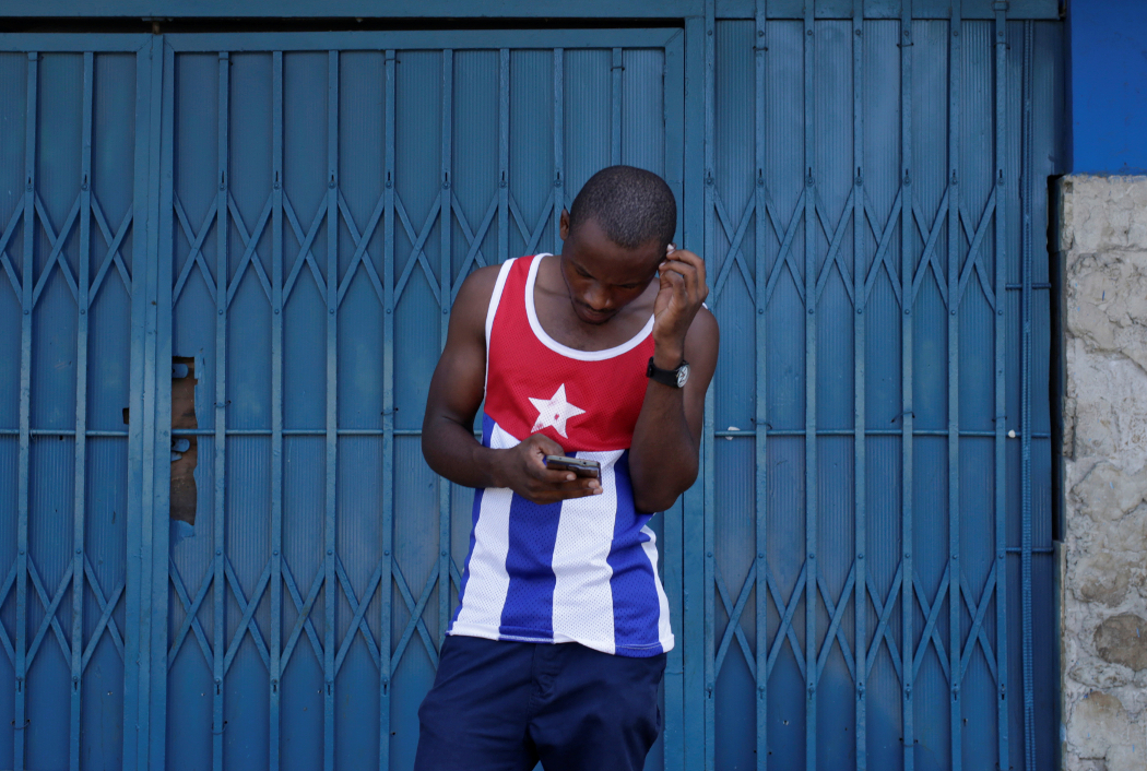 Jovem com camisa de Cuba e celular em frente a portão azul