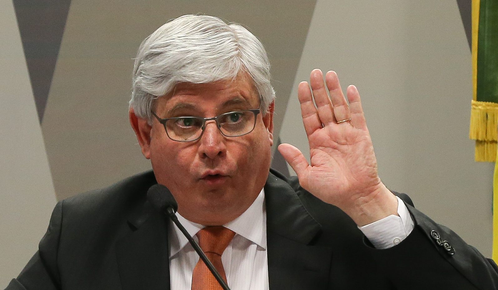 Político brasileiro em discurso, mão levantada, gesto enfático.