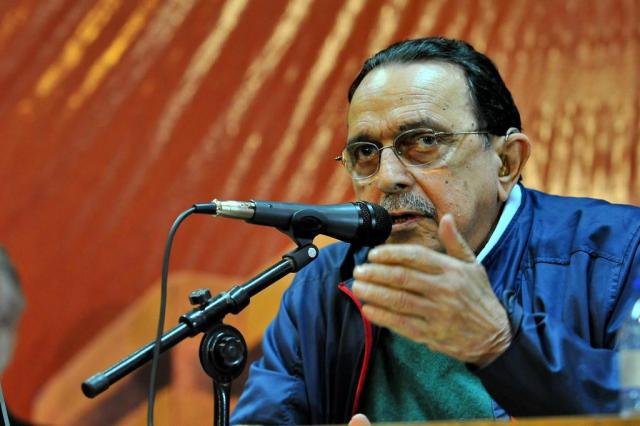 Homem discursando em evento com microfone e óculos.