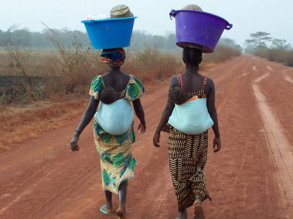 Mulheres com bebês e cargas caminhando em estrada de terra