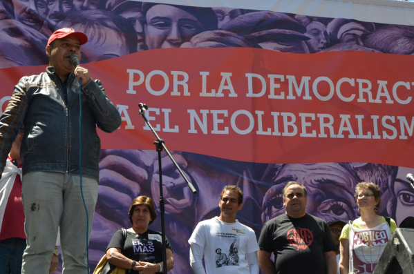 Homem discursa em palco: Por Democracia, Contra Neoliberalismo