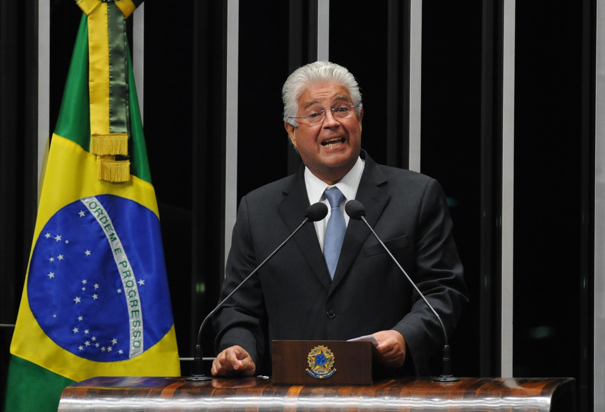 Político brasileiro discursa em evento oficial com bandeira nacional.