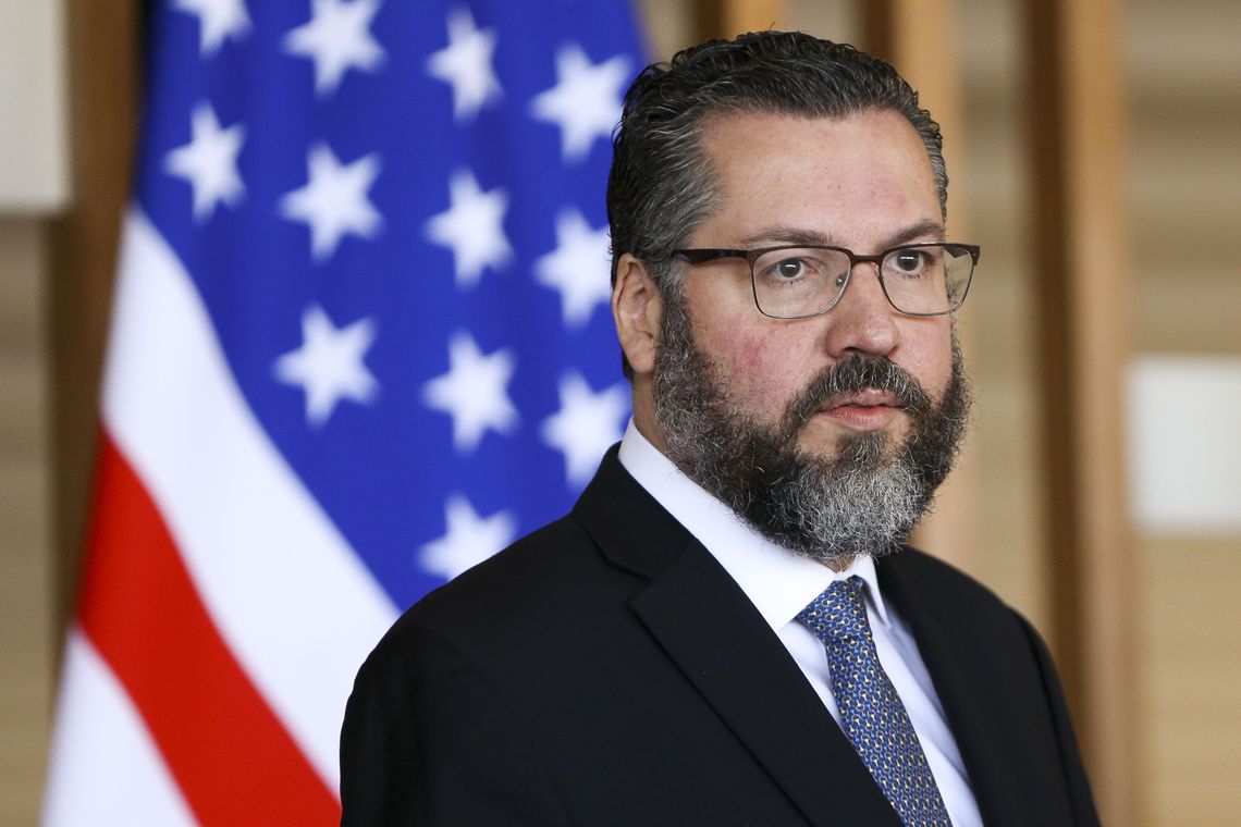 Homem barbudo de terno com óculos em frente à bandeira dos EUA.