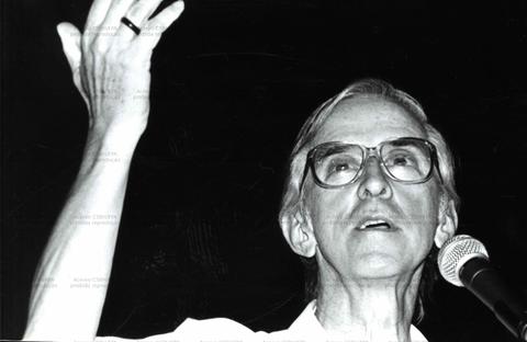 Homem discursando, mão levantada, óculos, em preto e branco.