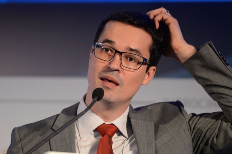 Homem pensativo com óculos, falando ao microfone em evento.