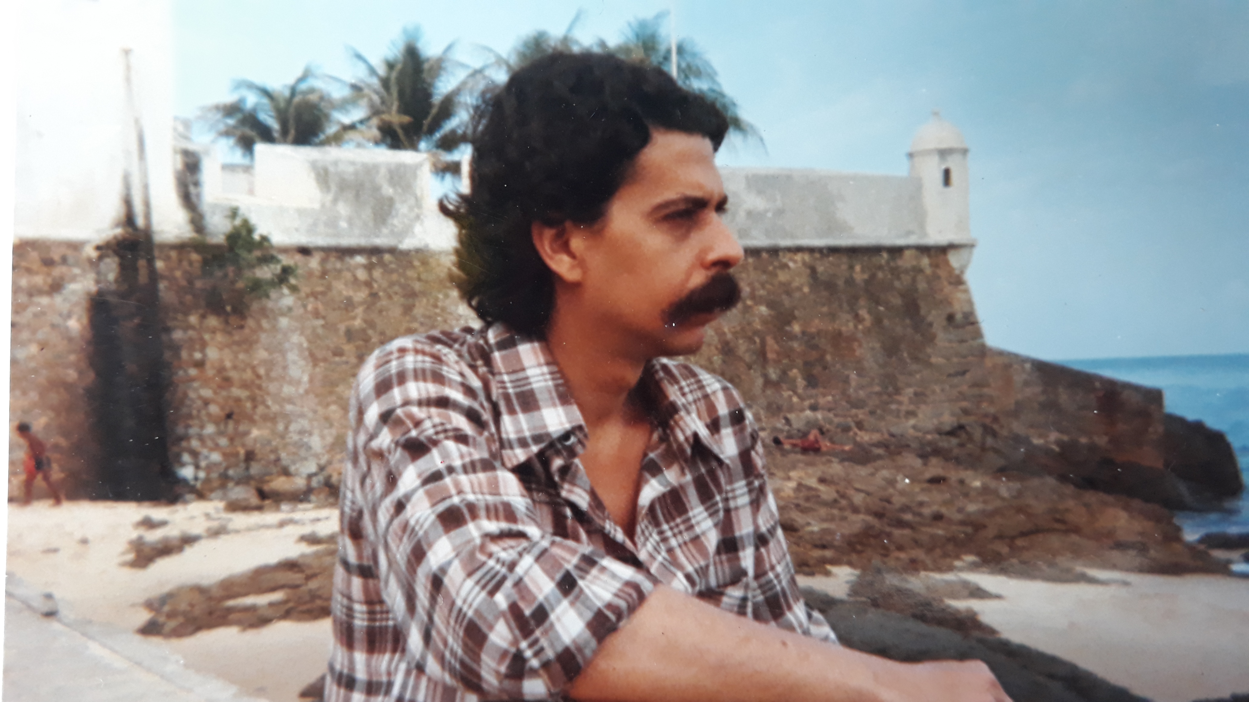 Homem pensativo em praia histórica com forte e coqueiros