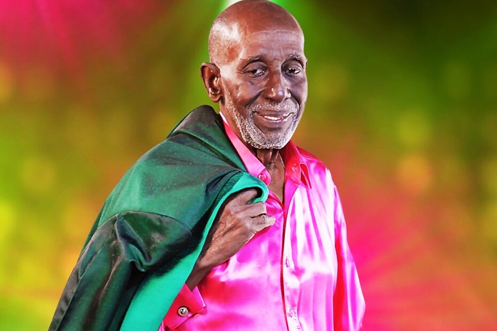 Homem Negro idoso sorridente, camisa rosa e jaqueta verde.