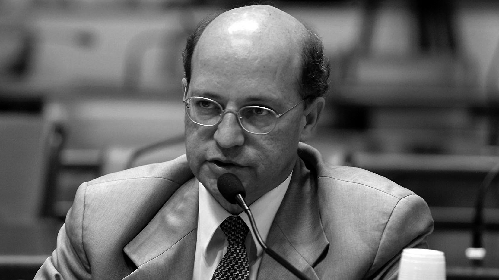 Homem de óculos discursa em evento formal, preto e branco