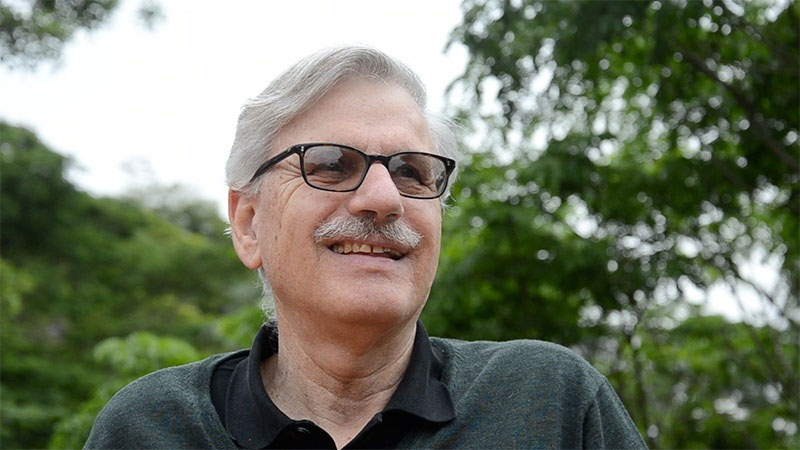 Homem sorridente com óculos e bigode em ambiente natural