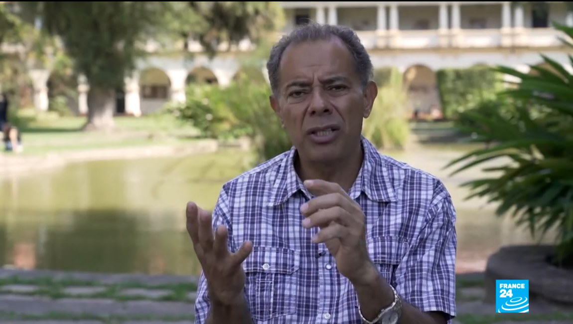 Homem em entrevista ao ar livre com fundo arquitetônico.