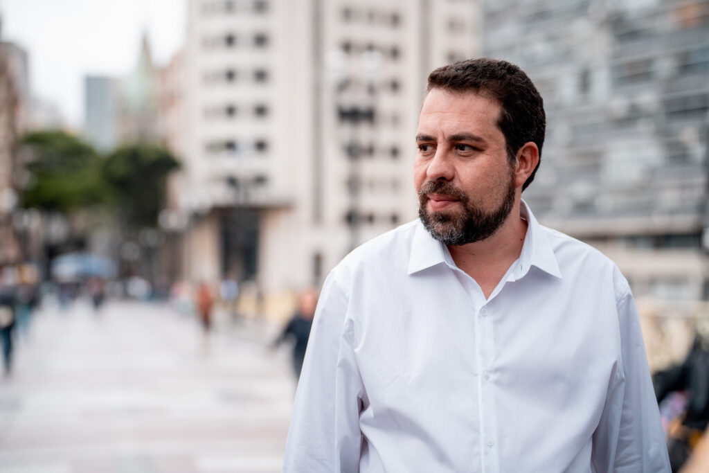 O deputado federal Guilherme Boulos veste camisa social branca e olha para direita. Ao fundo, em desfoque, o Viaduto do Chá
