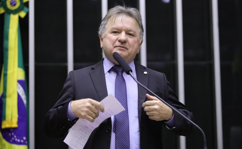 Político brasileiro discursando em plenário com bandeira nacional.