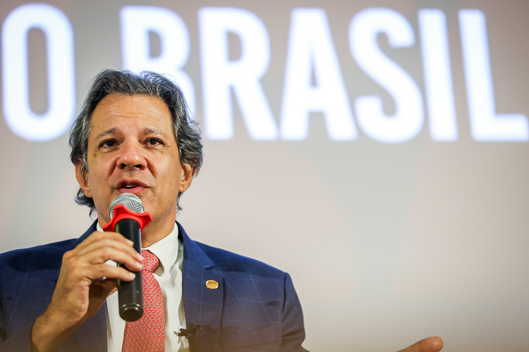 Fernando Haddad discursa em evento com "O Brasil" ao fundo.