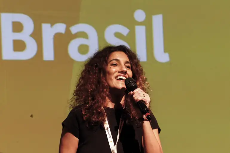 Mulher sorridente discursando no palco com "Brasil" ao fundo