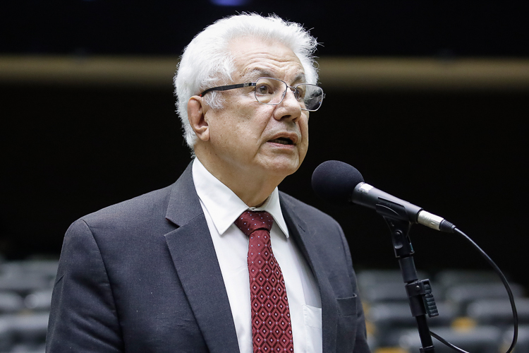 Homem idoso discursando em microfone, evento público.