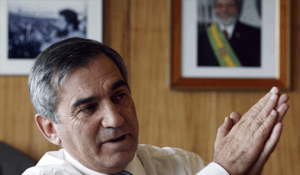 Homem em diálogo sério com retrato de Lula ao fundo