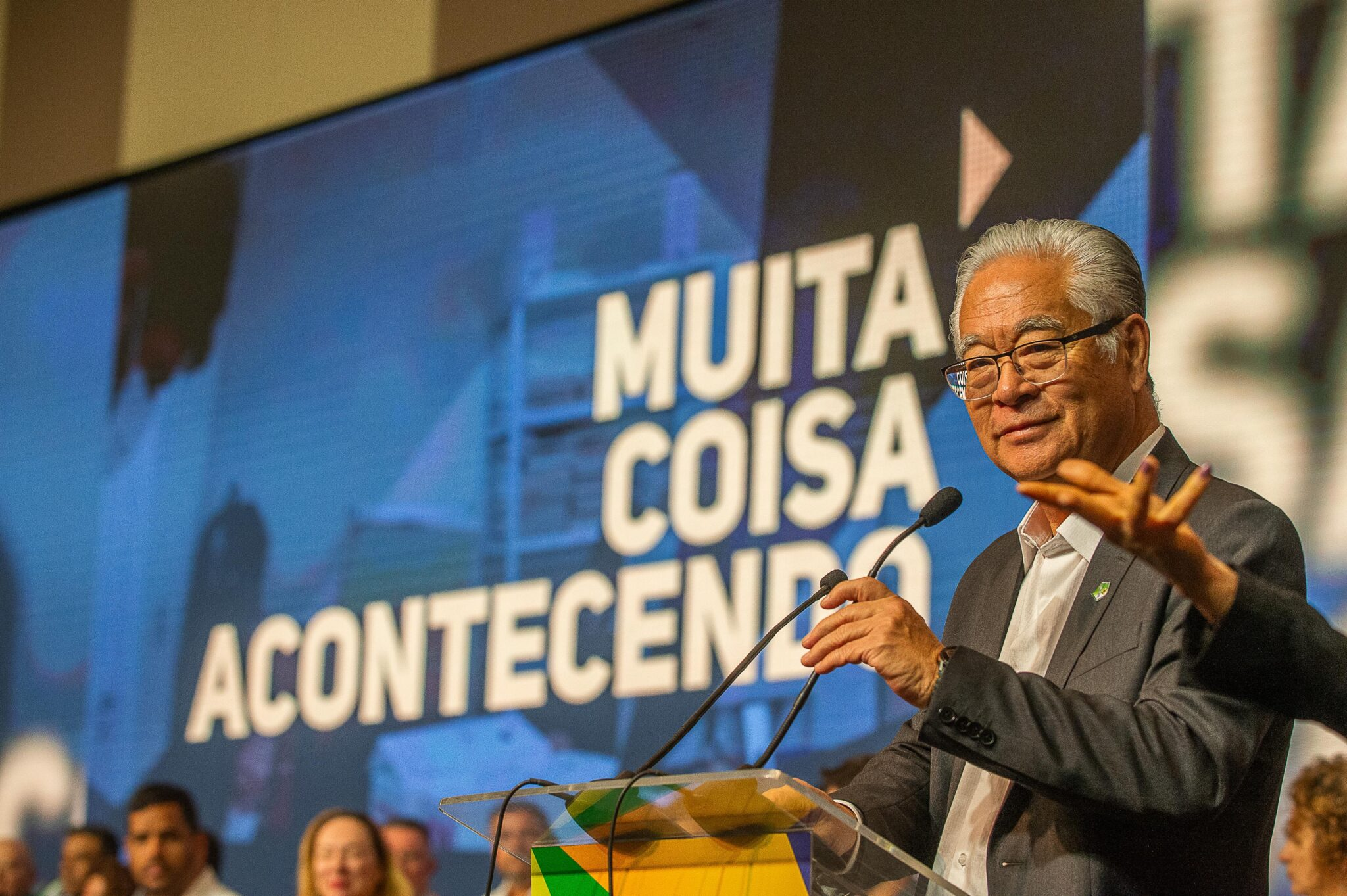 Homem sorridente discursando em evento público com tela ao fundo.