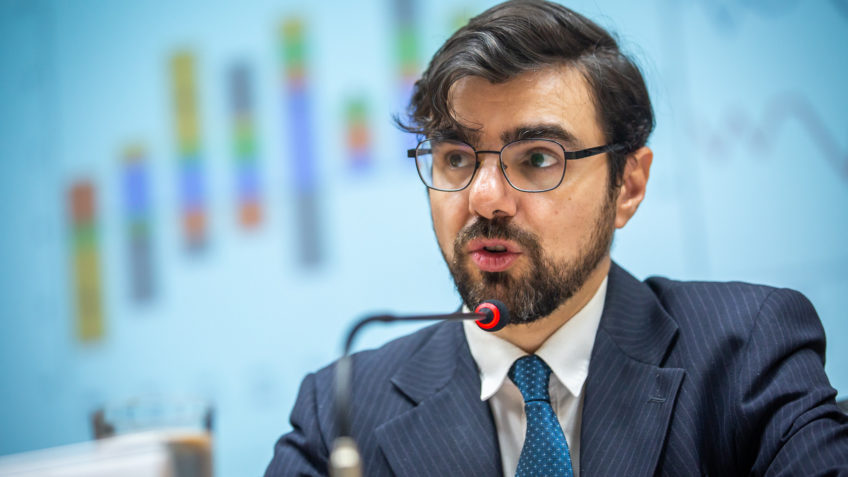 Homem de óculos em terno falando ao microfone com gráficos