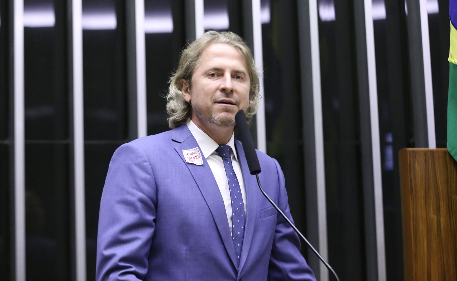 Homem discursando em plenário com broche 'AINDA ESTAMOS AQUI'